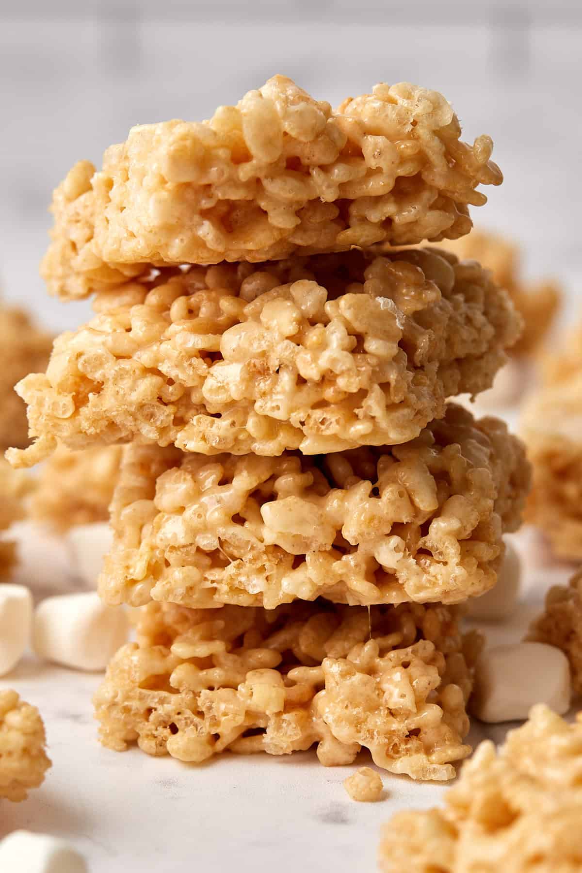 A close-up of four rice krispie treats stacked on top of each other, with mini marshmallows scattered around on a light surface. The treats appear gooey and golden brown.