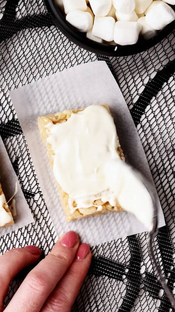 A hand holds a rice crispy treat on parchment paper while a spoon spreads white frosting on top; a bowl of mini marshmallows is visible above.