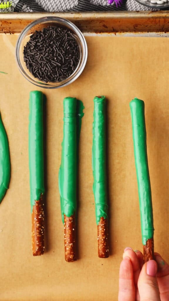 Four pretzel rods, partially dipped in green coating, are lined up on parchment paper with a bowl of chocolate sprinkles nearby.
