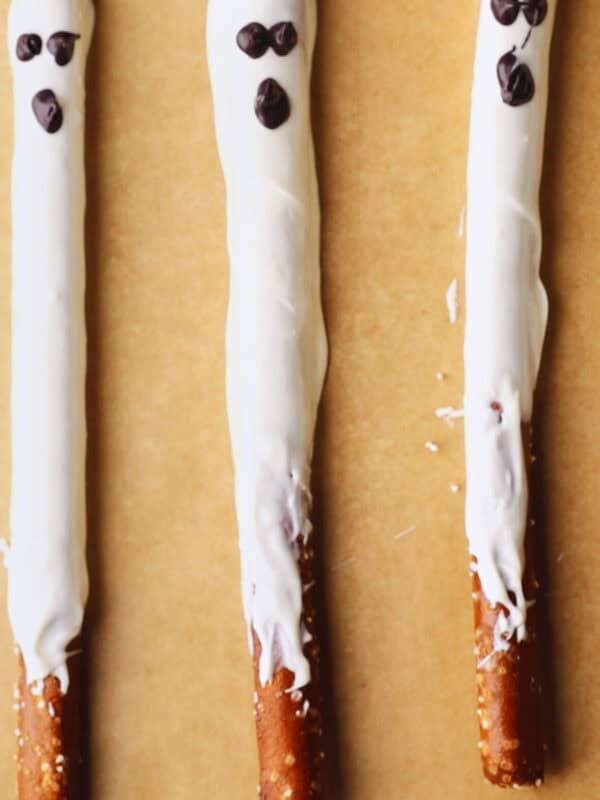 Three pretzel rods coated in white chocolate with chocolate  arranged as eyes and mouths, resembling ghost faces, placed on brown parchment paper.