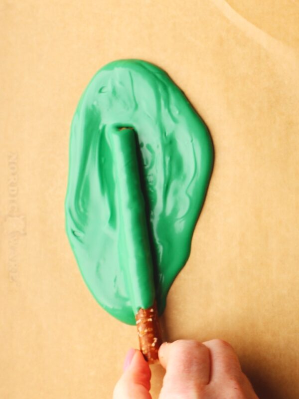 A hand holds a pretzel rod coated on one end with melted green candy, resting on parchment paper with a puddle of green candy.