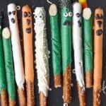 Assorted pretzel rods dipped in colorful candy coating, decorated with candy eyes and chocolate to resemble fun Halloween monsters and mummies, are arranged vertically on a black surface—perfect alongside your favorite halloween rice krispie treats.