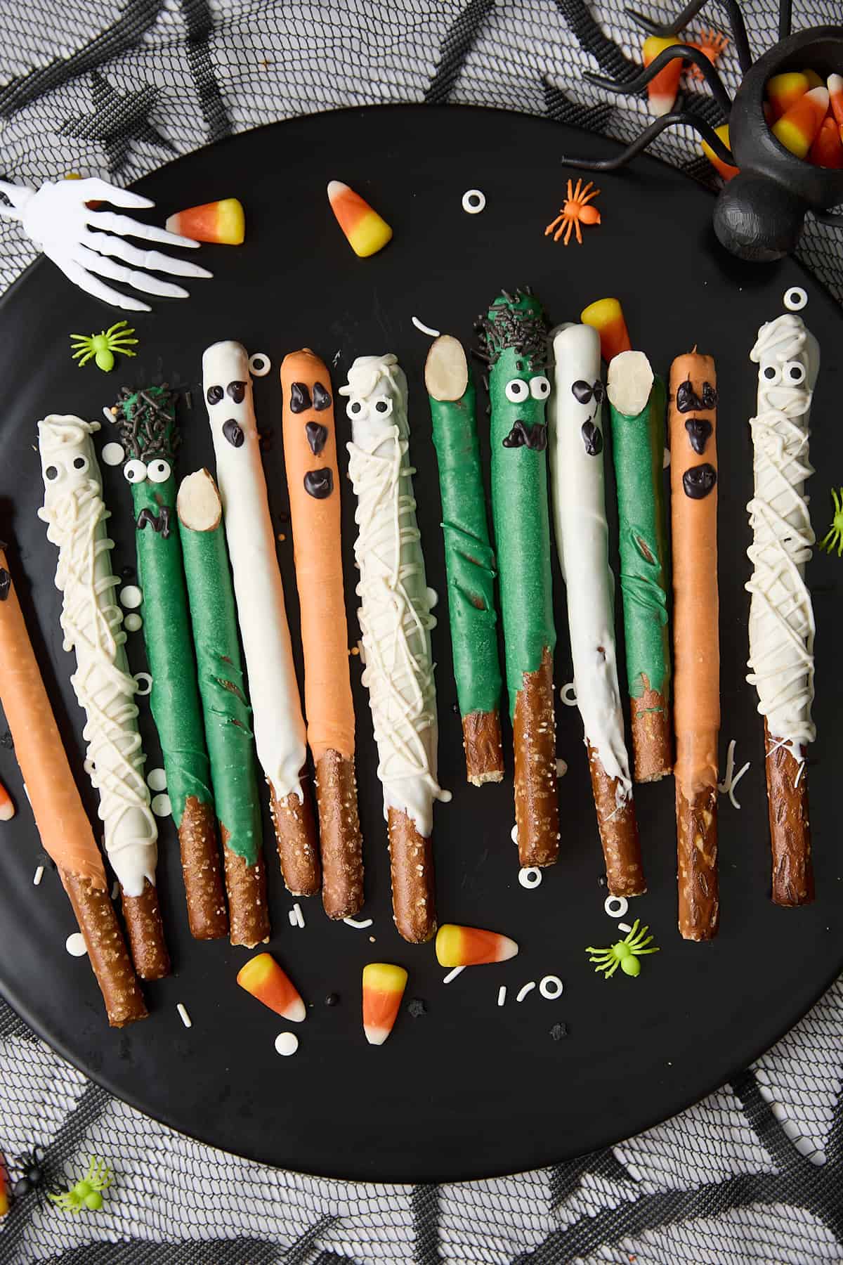 A platter of pretzel rods decorated as Halloween monsters, mummies, and ghosts with white, green, and orange candy coatings and candy eyes, surrounded by candy corn and Halloween decorations.