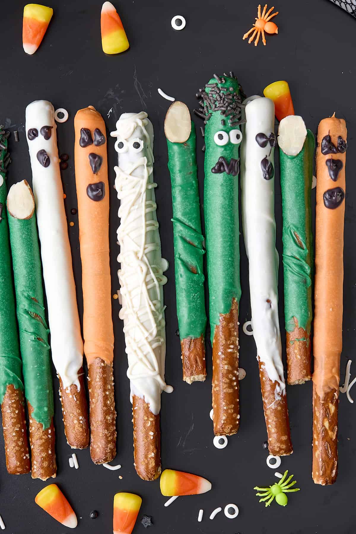 Pretzel rods decorated as Halloween monsters with colorful candy coating, candy eyes, and sprinkles, arranged on a black surface with candy corn, toy spiders, and sprinkles scattered around.