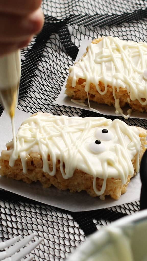 A hand decorates a Halloween Rice Krispies treat with white icing to resemble a mummy, complete with candy eyes, on parchment paper. Another Halloween Rice Krispies treats creation sits in the background on a black netted surface.