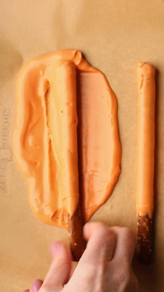A hand presses a pretzel rod into a puddle of melted orange candy on parchment paper, next to another pretzel rod coated evenly with melted orange candy.