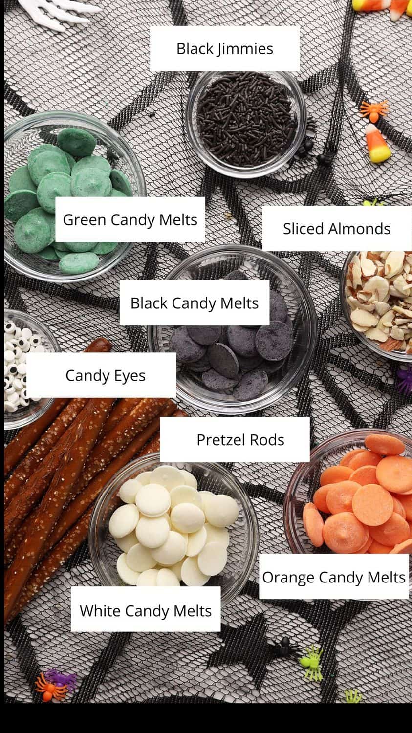 An overhead view of bowls containing black jimmies, green, black, white, and orange candy melts, sliced almonds, candy eyes, and pretzel rods, all labeled and arranged on a gray patterned cloth.