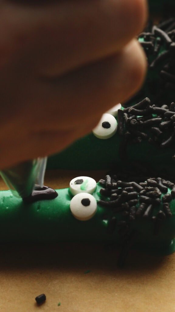 A close-up of a hand decorating a green, monster-themed Halloween Rice Krispies Treats recipe with candy eyes, black sprinkles for hair, and black icing for a mouth.
