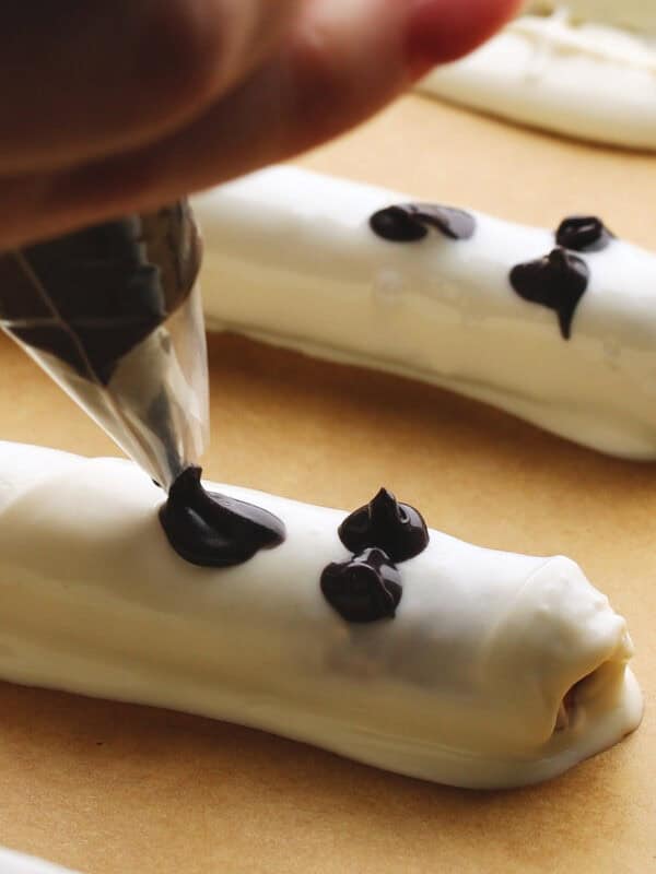 A hand uses a piping bag to add small dots of dark chocolate onto white chocolate-covered pretzel rods on a sheet of parchment paper.