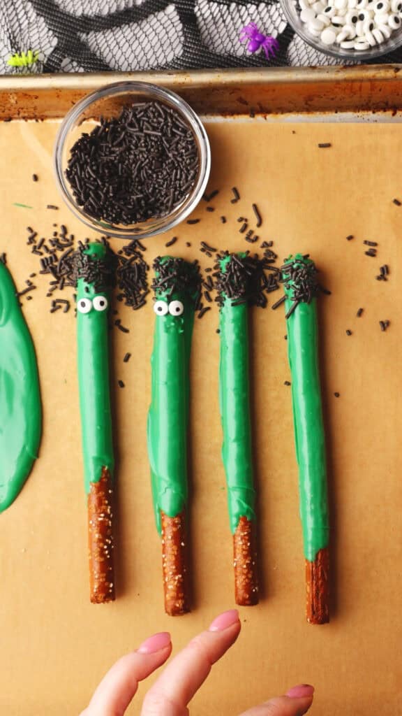 Four pretzel rods dipped in green candy coating with candy eyes and black sprinkles, arranged like monsters on parchment—perfect for serving alongside your favorite halloween rice krispie treats at your spooky party.