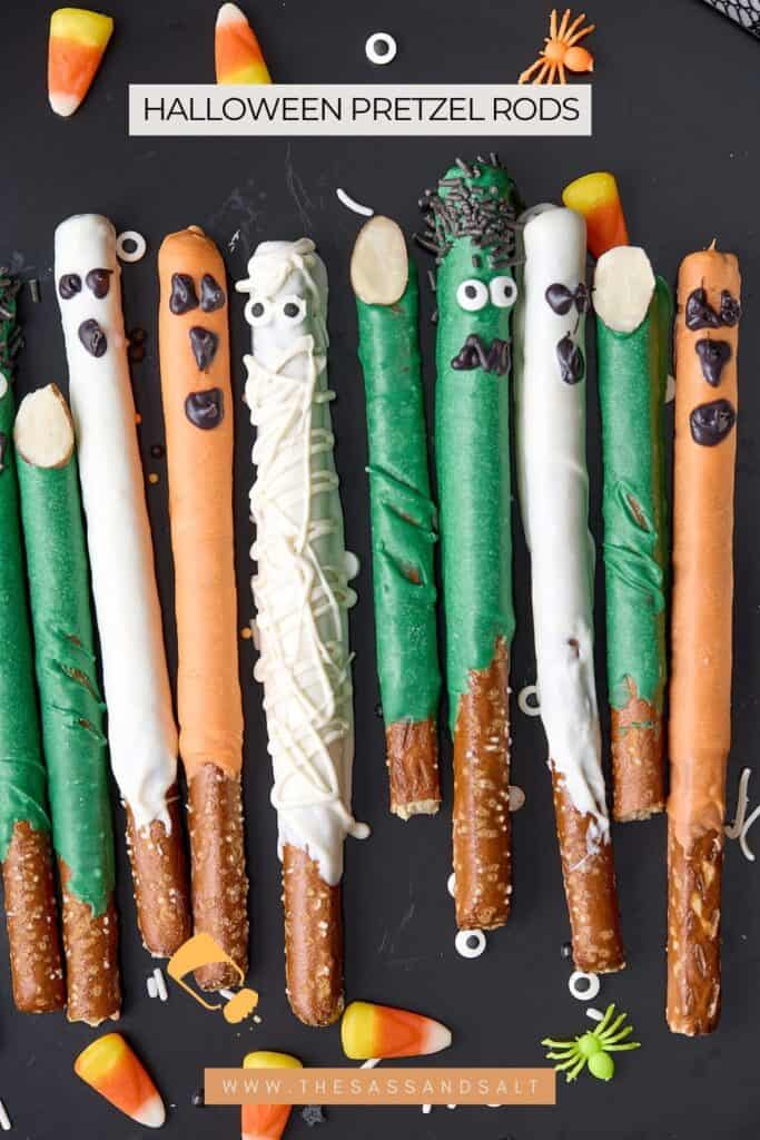 A row of pretzel rods dipped in white, orange, and green chocolate, decorated as Halloween monsters and mummies, with candy eyes and icing faces. Candy corn and festive sprinkles are scattered around.