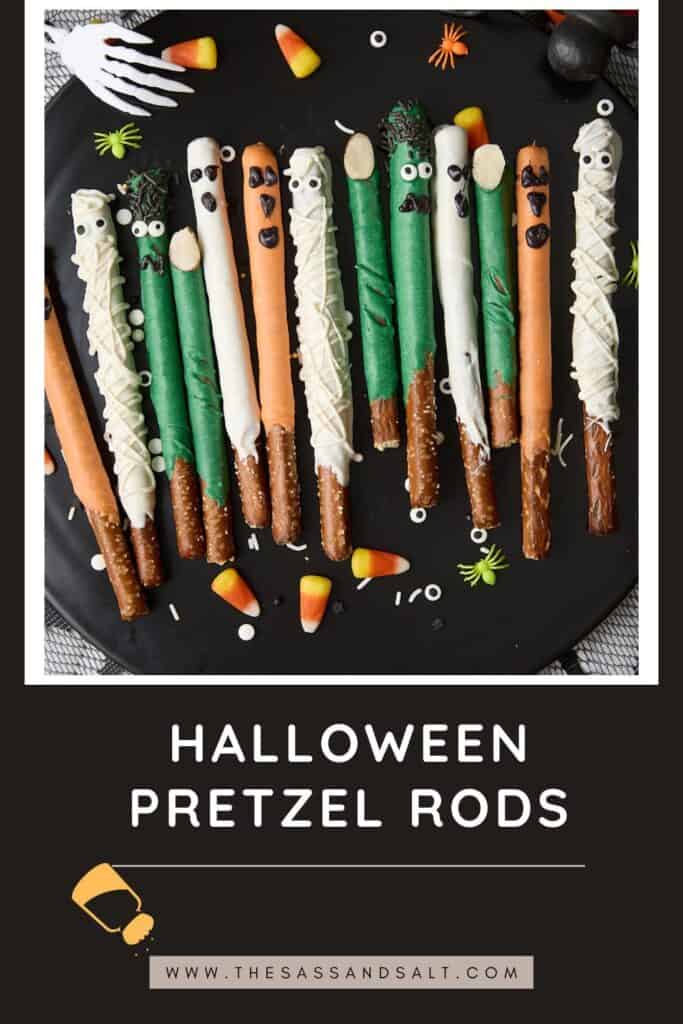 A plate of Halloween-themed pretzel rods decorated as mummies, monsters, and pumpkins with colorful candy melts, candy eyes, and Halloween sprinkles. Candy corn and fake spiders are scattered around. Text reads "Halloween Pretzel Rods.