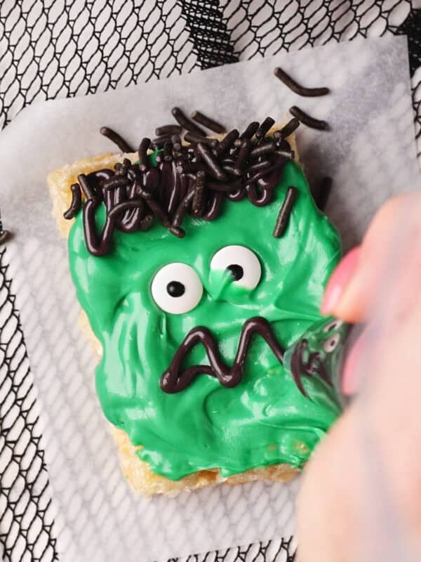 A hand decorates a Halloween Rice Krispie Treat to resemble a Frankenstein monster, using green frosting, candy eyes, black icing for the mouth, and chocolate sprinkles for hair. The treat rests on parchment paper over a mesh surface.