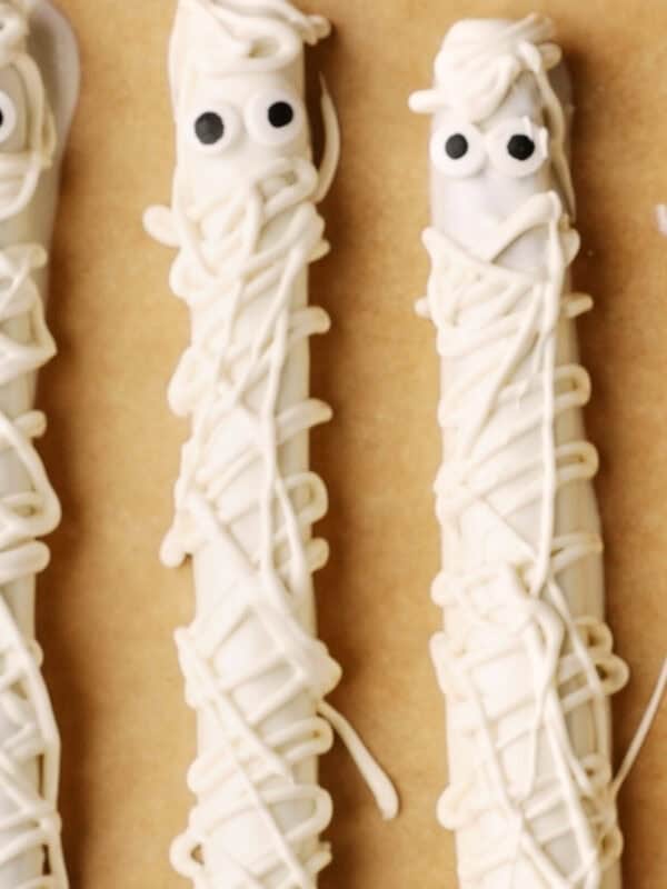 Two pretzel sticks decorated to look like mummies, covered in white chocolate with drizzled white lines and topped with candy eyes, on a brown surface.