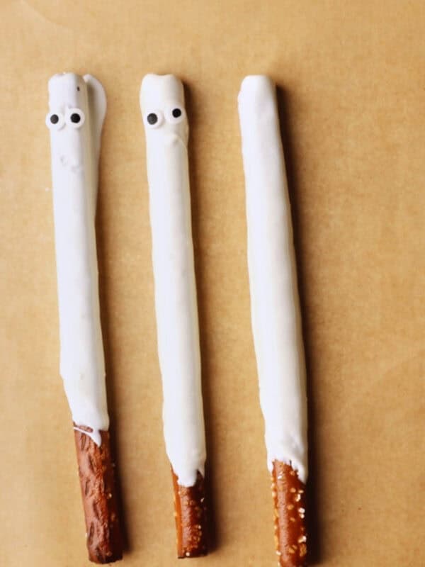 Three pretzel rods partly dipped in white chocolate, arranged vertically on brown parchment paper. Two have candy eyes, resembling ghost faces, while the third is plain.