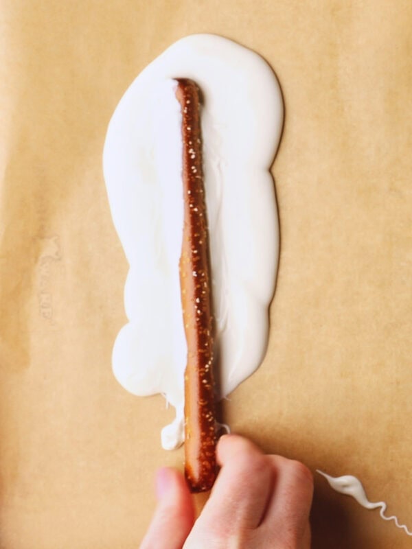 A hand dipping a pretzel rod into melted white chocolate, spreading it on a sheet of parchment paper.