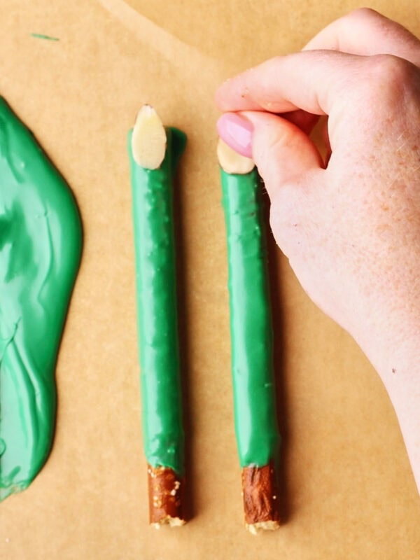 A hand is placing an almond slice on the tip of a pretzel stick coated in green candy, next to another similar pretzel on parchment paper. Melted green candy is smeared on the left side.