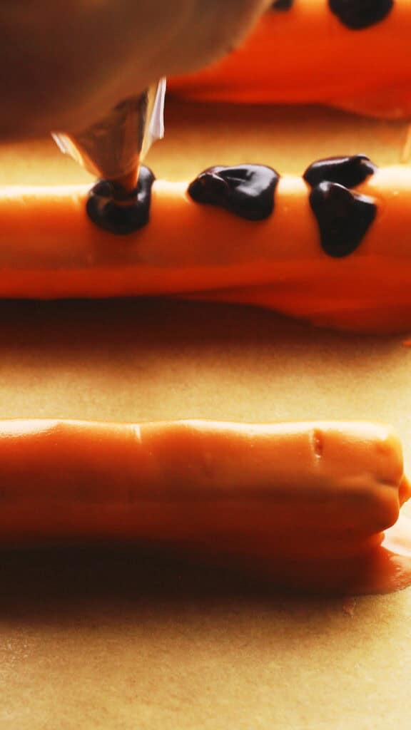 Orange-coated pretzel sticks on parchment paper, with someone piping small black chocolate eyes nose and mouth to resemble a pumpkin.