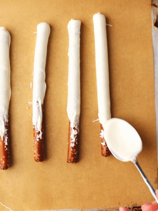 Four pretzel rods coated with white chocolate are lined up on parchment paper, while a hand uses a spoon to drizzle more melted white chocolate onto one of the pretzels.