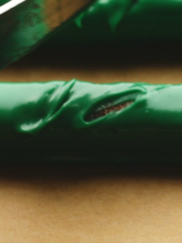 A close-up of a green-coated pretzel rod cut with a knife on a brown surface, showing a tear in the green coating revealing the pretzel rod underneath.