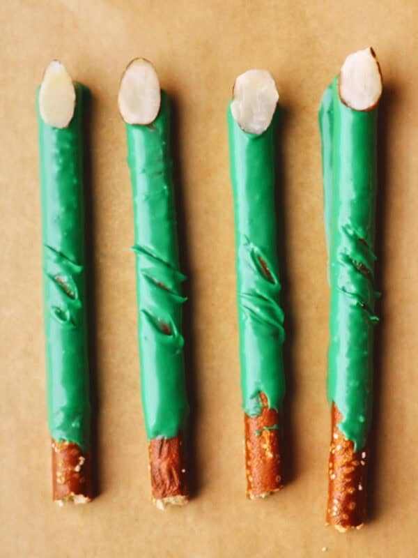 Four pretzel sticks partially coated in green candy, with almond slices on the tips, resembling witch fingers, are arranged in a row on brown parchment paper.