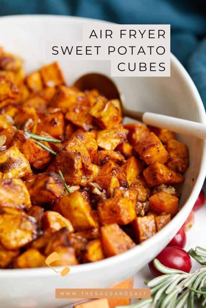 A bowl filled with golden-brown air fryer sweet potato cubes, garnished with herbs, sits on a table. A spoon rests in the bowl, and cranberries are scattered like a festive brownie graveyard. Text overlay reads, "Air Fryer Sweet Potato Cubes.