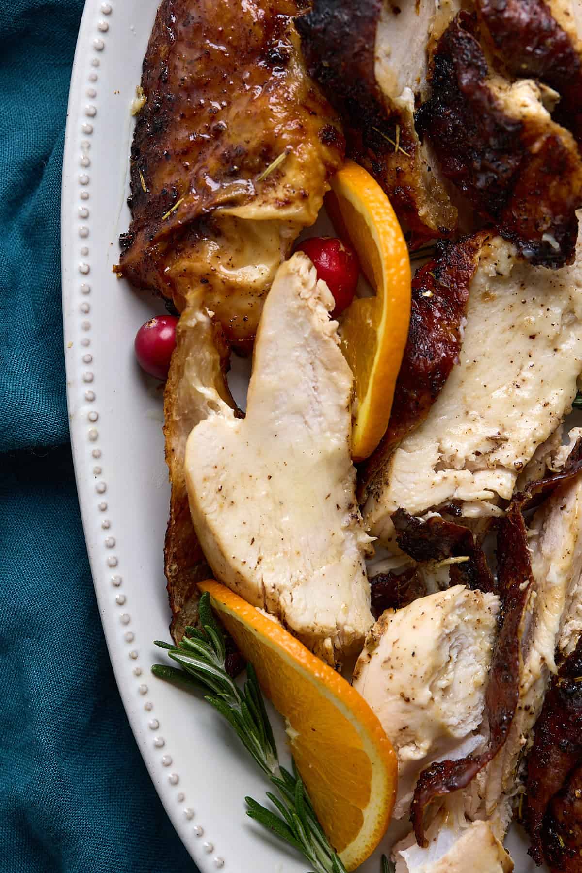 Sliced roasted Air Fryer Turkey Breast with crispy skin, garnished with orange slices, cranberries, and rosemary, served on a white beaded plate atop a teal cloth.