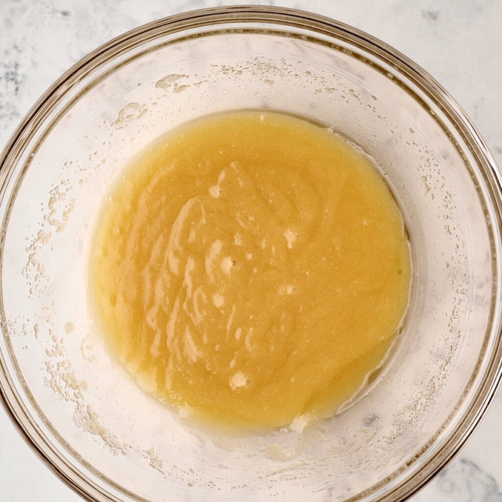 A clear glass bowl filled with a smooth, light yellow mixture sits on a white, lightly speckled countertop.