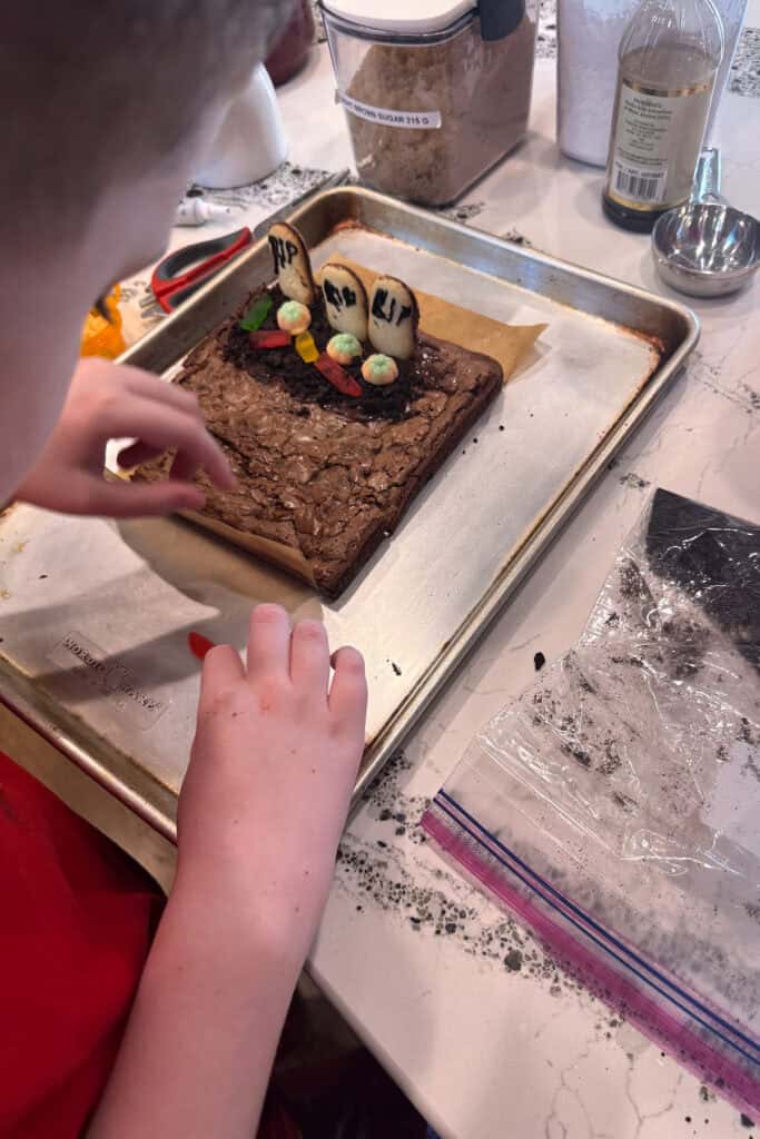 A child decorates a brownie graveyard with cookie tombstones, colorful candies, and icing on a baking sheet, surrounded by baking ingredients on a kitchen counter.
