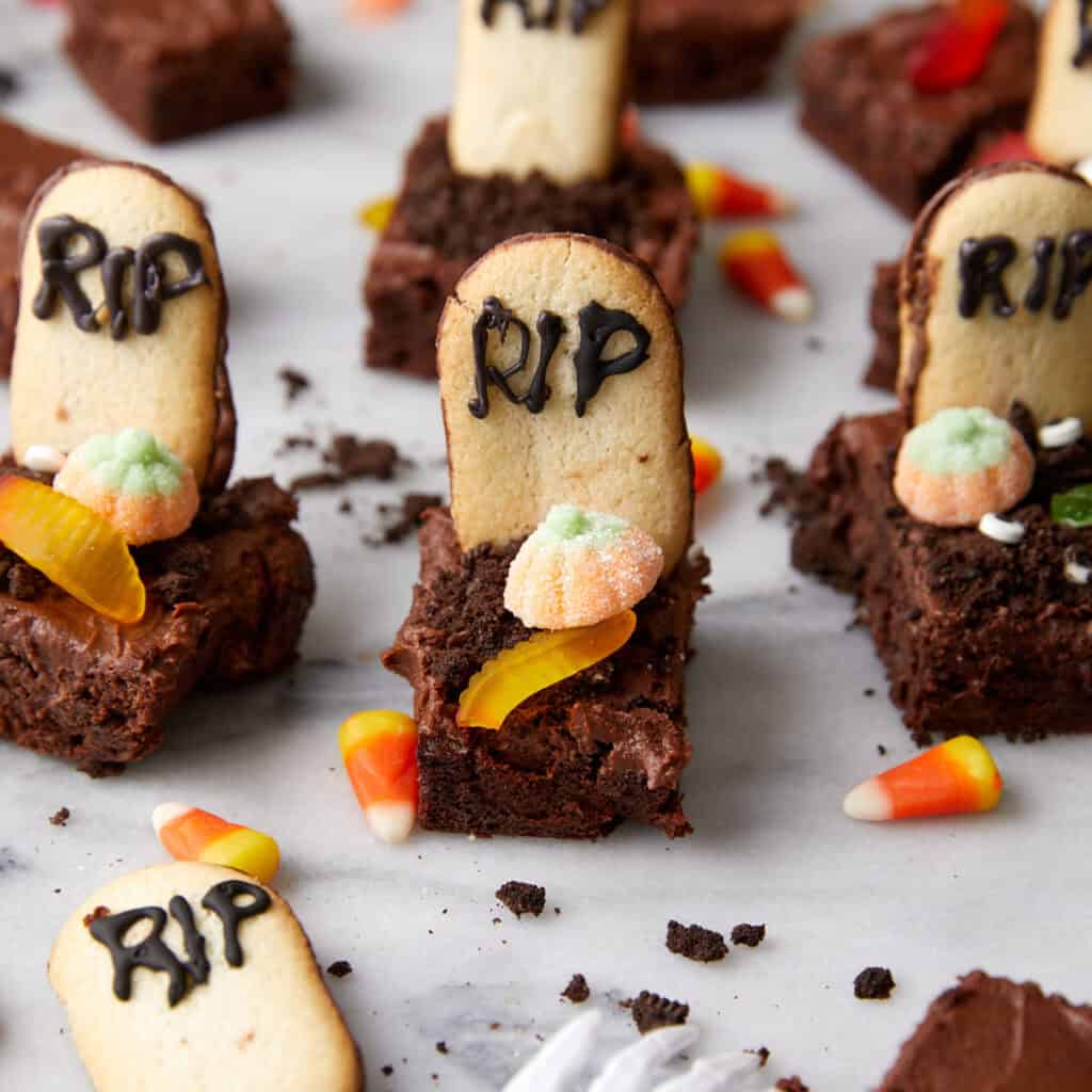 Brownie squares decorated as Halloween graves with cookie "tombstones" labeled "RIP" in icing, gummy worms, candy pumpkins, and candy corn accents on a marble surface.