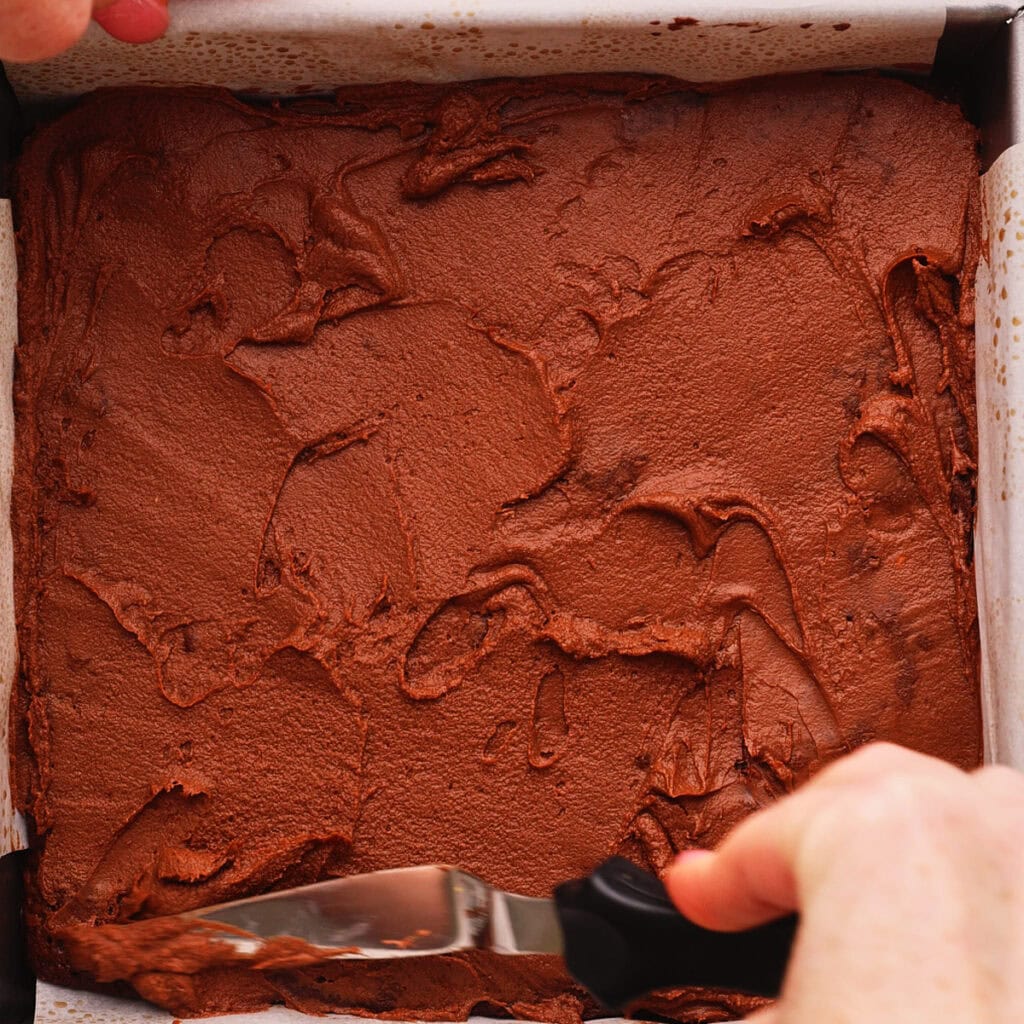 A close-up of hands spreading chocolate frosting evenly over a square cake in a parchment-lined baking pan using an offset spatula.