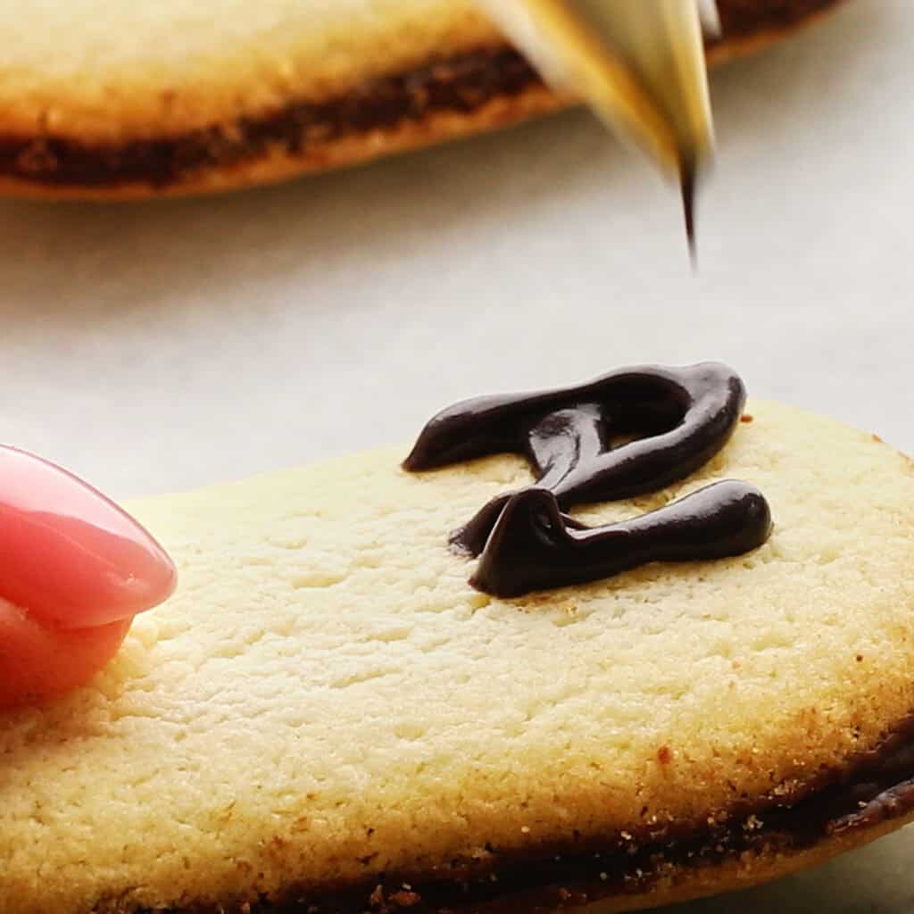 A hand with pink nails holds a cookie while chocolate icing is being piped onto it in the shape of the letter "RIP".