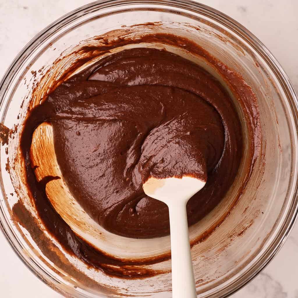 A glass bowl filled with thick chocolate batter being mixed with a white spatula, placed on a light marble surface.