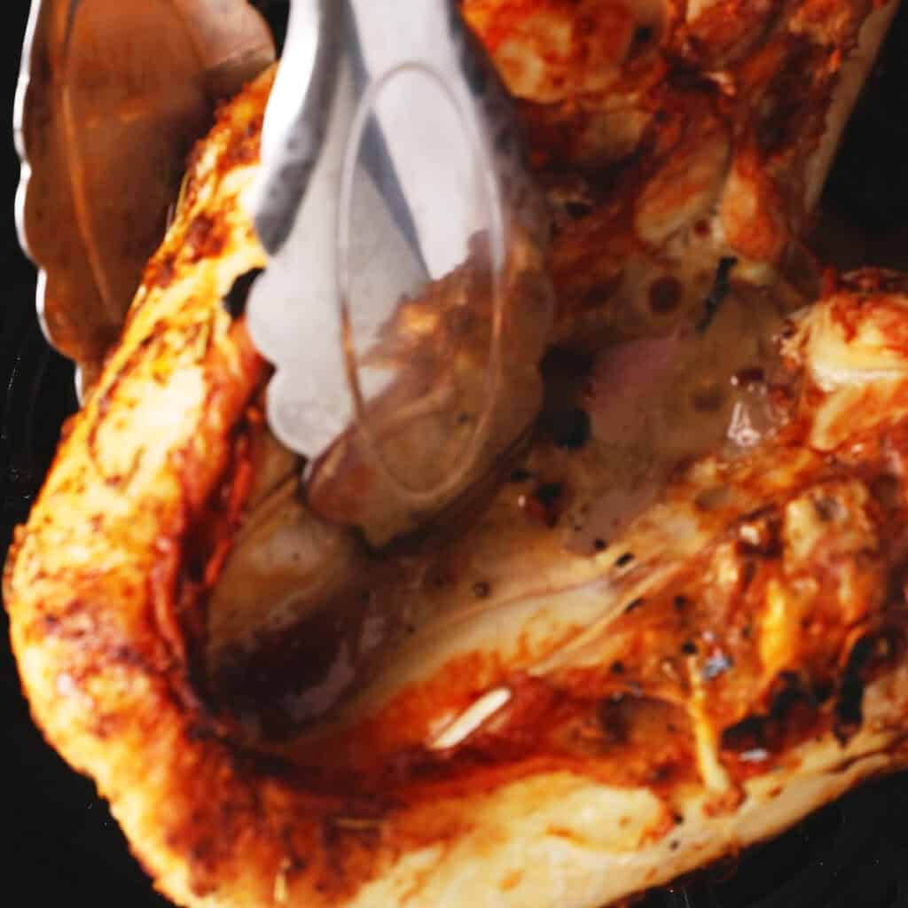 A close-up of metal tongs holding a piece of grilled, seasoned  turkey breast in air fryer, showcasing its golden-brown, crispy exterior.