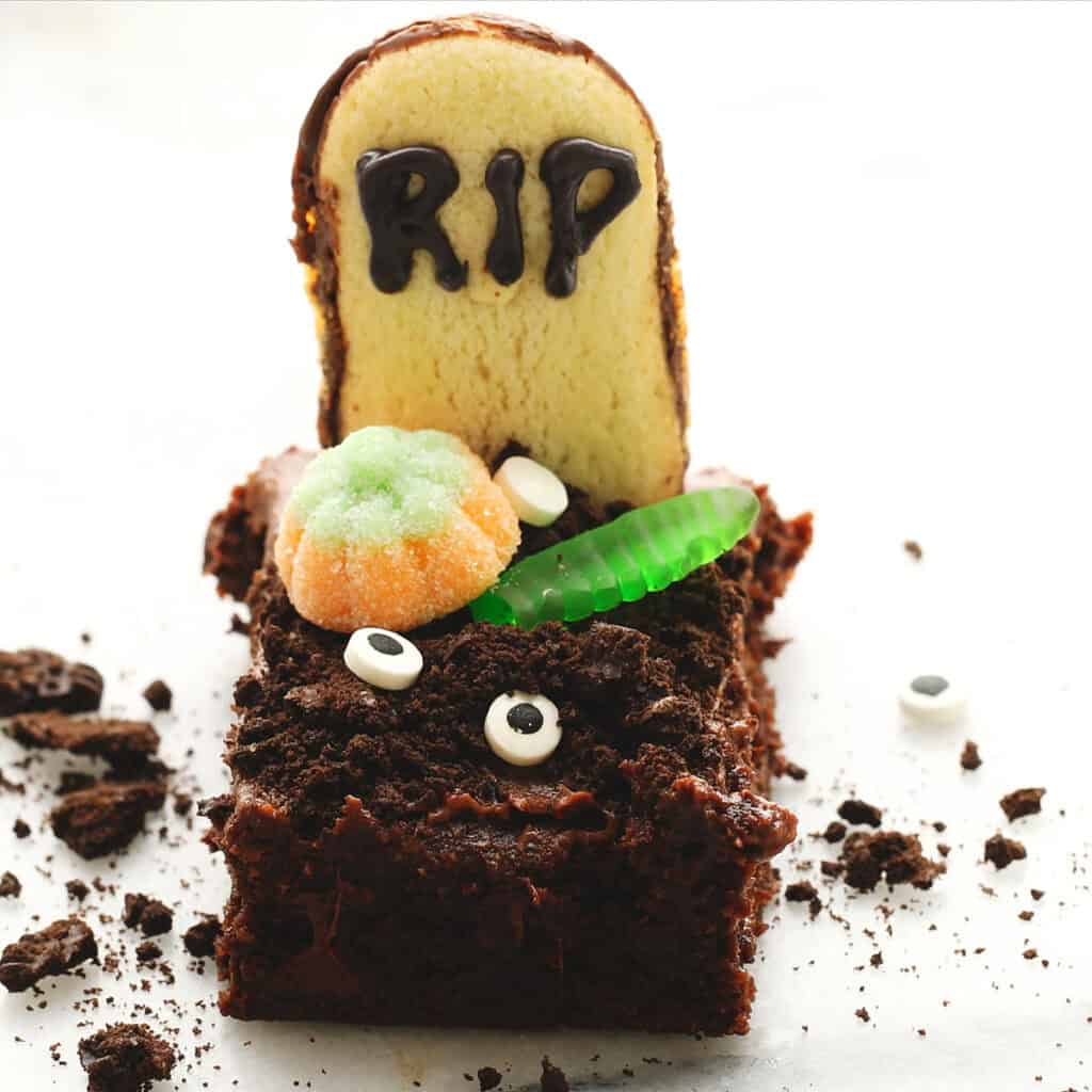 A Halloween-themed brownie decorated to look like a grave, topped with crushed cookies, a cookie tombstone with "RIP" written in chocolate, a gummy worm, candy pumpkin, and candy eyes.