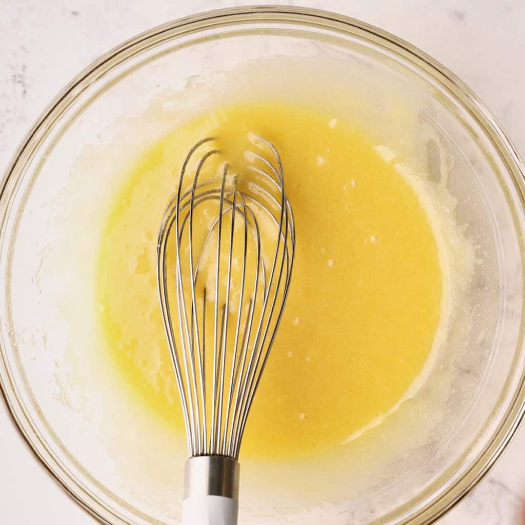 A metal whisk rests in a glass bowl filled with a smooth, yellow batter mixture, set on a light countertop.