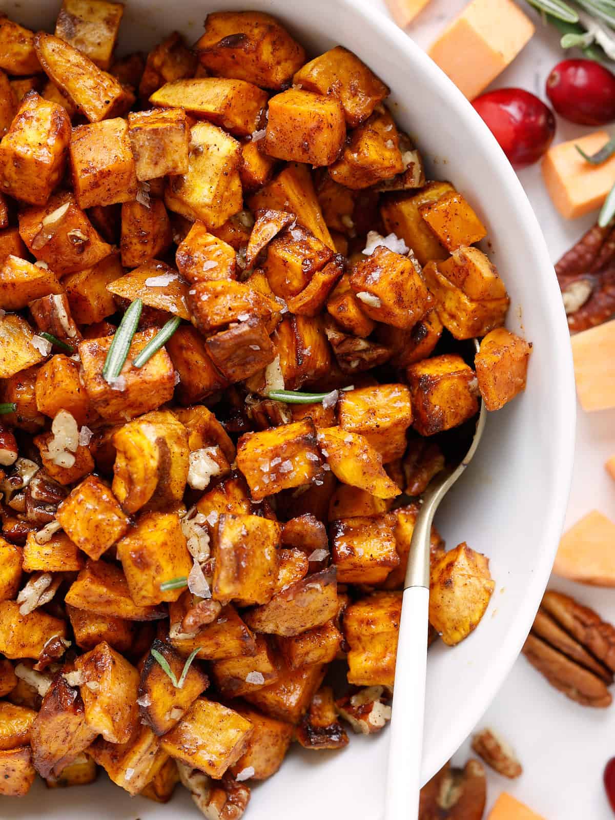 A close-up of a bowl filled with roasted sweet potato cubes, garnished with chopped pecans, rosemary, and spices. A spoon rests in the bowl, and raw sweet potatoes, cranberries, and pecans are scattered nearby. The rosemary is just for styling and not part of the recipe.