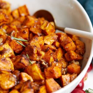 A close-up of air fryer sweet potato cubes in a white dish, garnished with herbs and a spoon resting inside. The cubed sweet potatoes have a golden-brown, caramelized appearance.