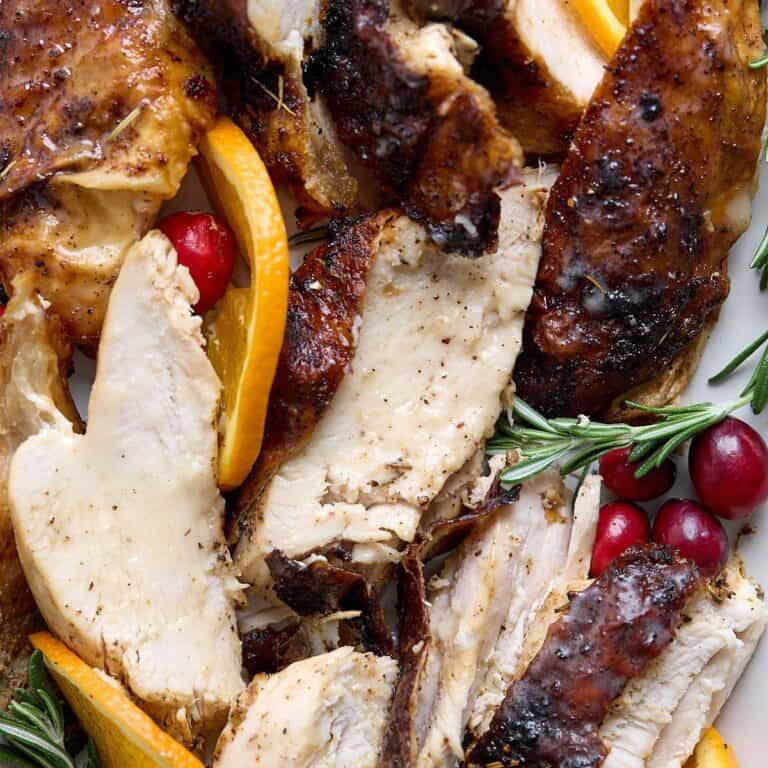 Sliced roasted turkey breast with crispy browned skin, garnished with fresh rosemary, orange slices, and cranberries on a white plate.