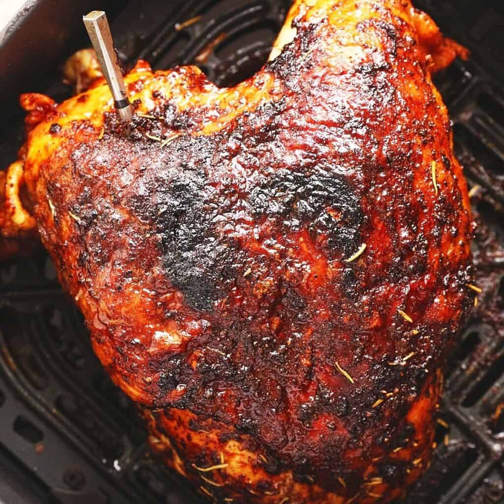 A close-up of Air Fryer Turkey Breast with a meat thermometer inserted, resting on a dark air fryer basket. The golden-brown, crispy skin is seasoned with visible herbs and charred spots for added flavor.