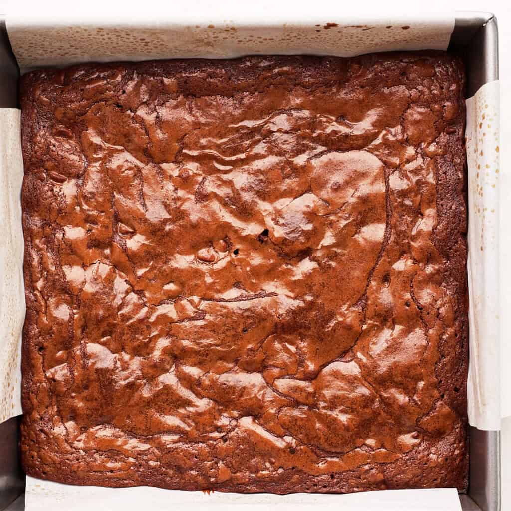 A freshly baked pan of brownies with a shiny, cracked top, still in a parchment-lined square baking dish.