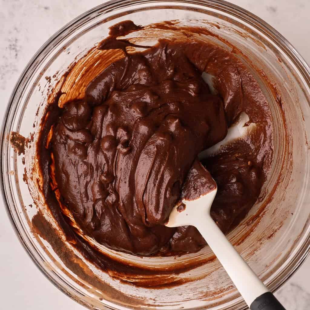 A glass bowl filled with thick, glossy chocolate brownie batter being mixed with a white spatula on a light-colored surface.