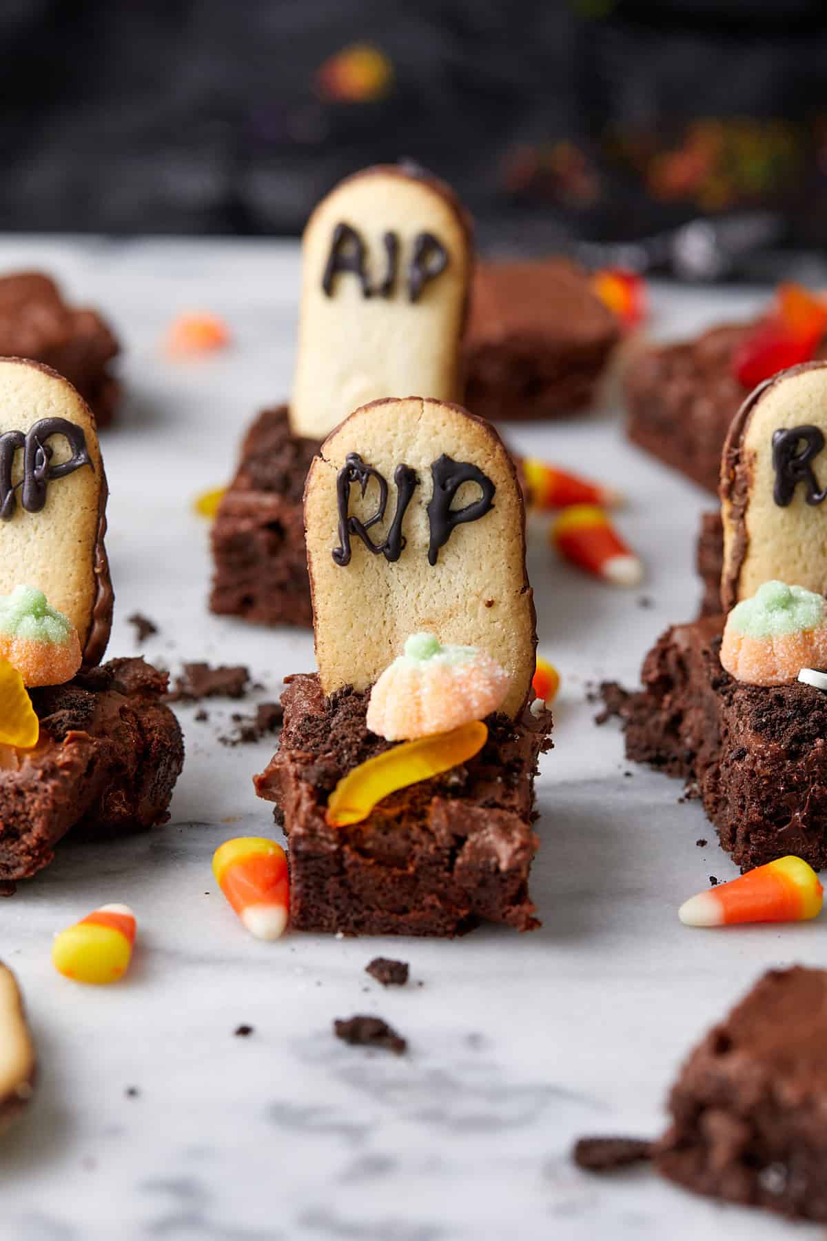 A close-up of Halloween-themed brownies topped with cookie "tombstones" labeled "RIP," candy pumpkins, gummy worms, and candy corn, arranged on a marble surface.