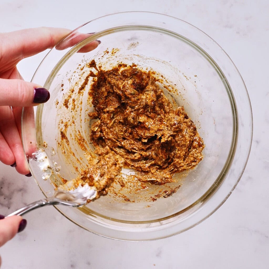 A person holding a glass bowl of thick, butter and spice mixture, stirring it with a spoon on a white marble surface