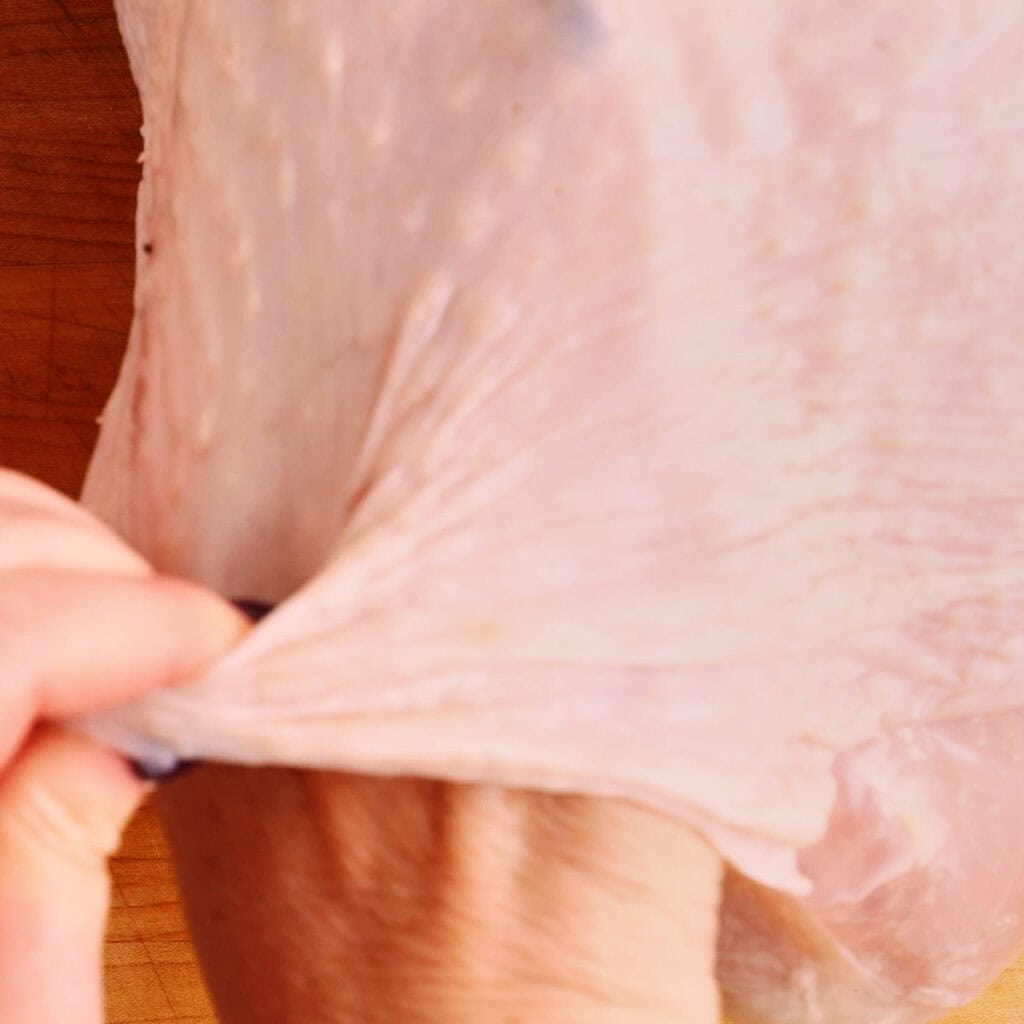 A hand pulls back the skin from a raw turkey breast on a wooden surface, prepping it for an Air Fryer Turkey Breast recipe by showing the separation between the skin and meat.