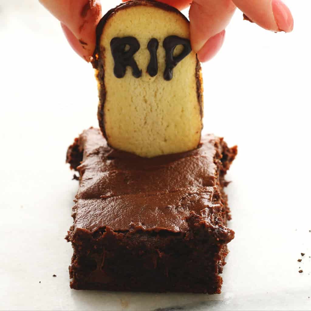 A hand places a cookie decorated with "RIP" in black icing onto a rectangular chocolate frosted brownie, resembling a tombstone in a grave.