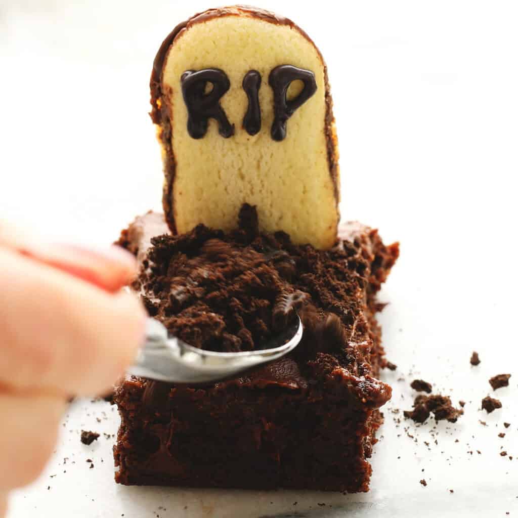 A hand uses a spoon to sprinkle crushed chocolate cookies onto a chocolate cake decorated with a cookie shaped like a tombstone, iced with the letters “RIP.”