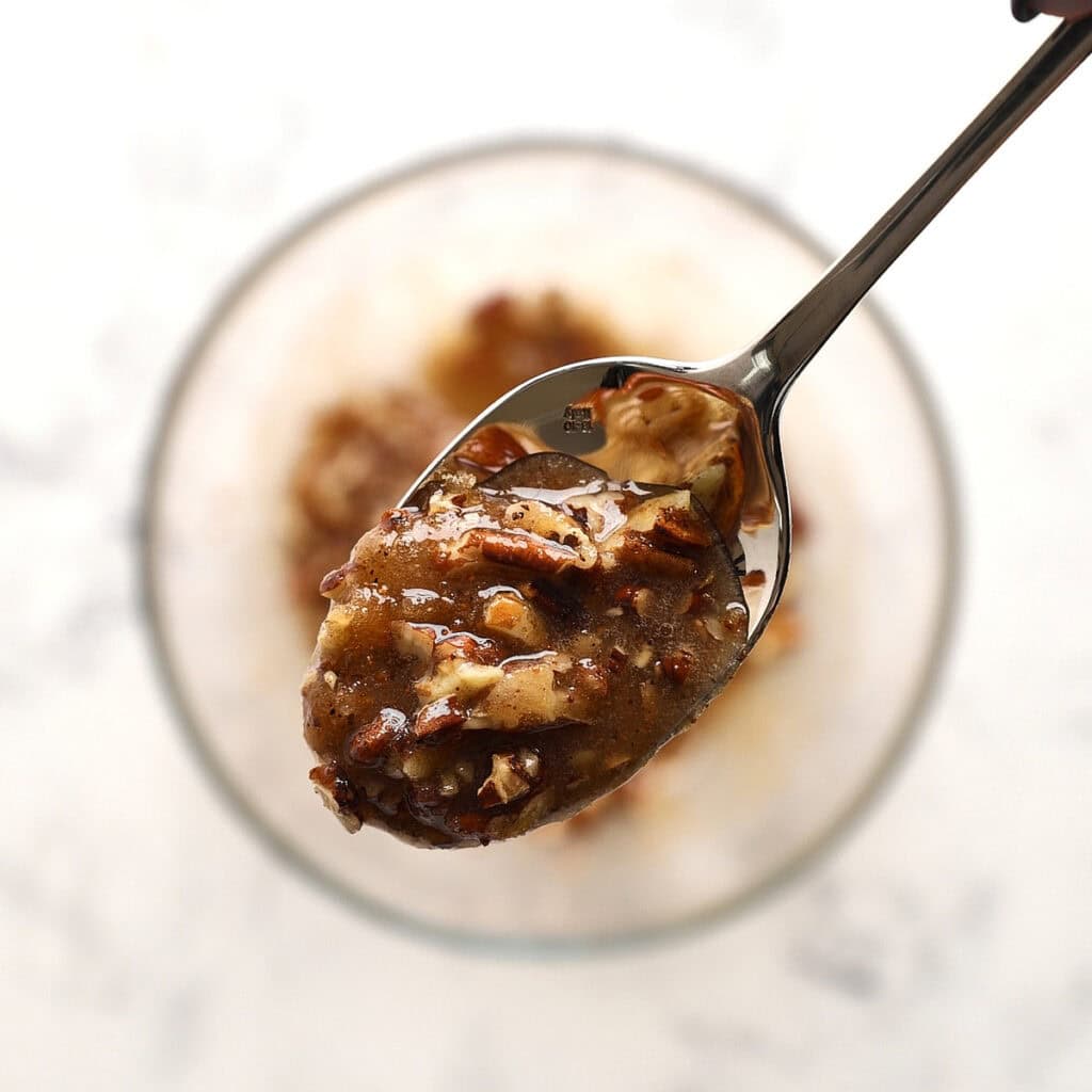 A close-up of a spoon holding a mixture of chopped nuts, butter, cinnamon and brown sugar held above a glass bowl containing more of the same mixture. The background is light and out of focus.
