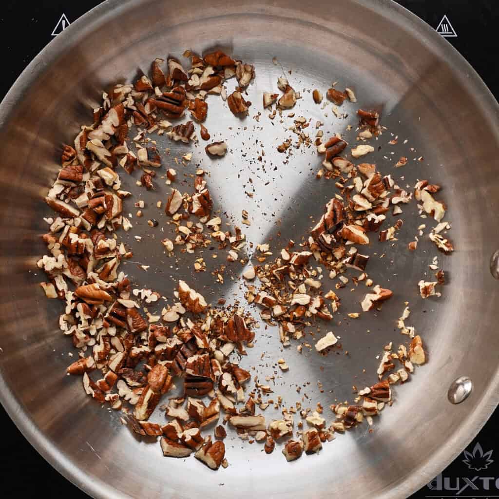 Chopped pecans are being toasted in a stainless steel skillet on a stovetop, with the nuts scattered mostly around the edges of the pan.