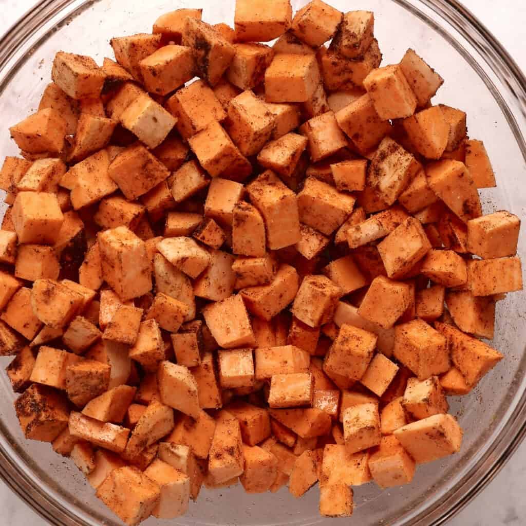 A glass bowl filled with small, evenly cut sweet potato cubes coated in spices, ready to be cooked.